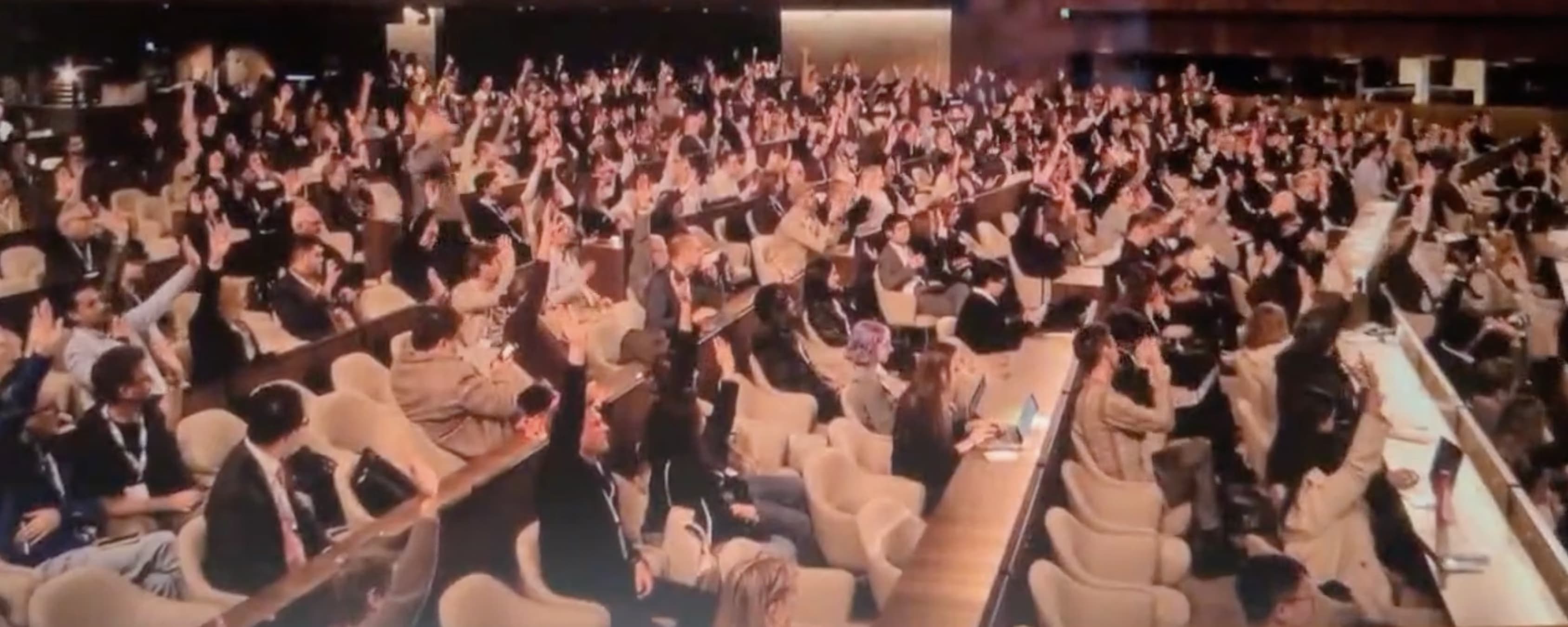 An auditorium filled with conference attendees, with the vast majority raising their hands in a show of support.
