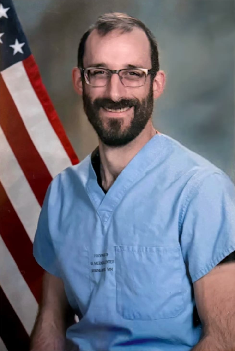 A smiling man with a beard and glasses, wearing light blue medical scrubs with "VA MEDICAL CENTER" visible on the chest, posing in front of an American flag.