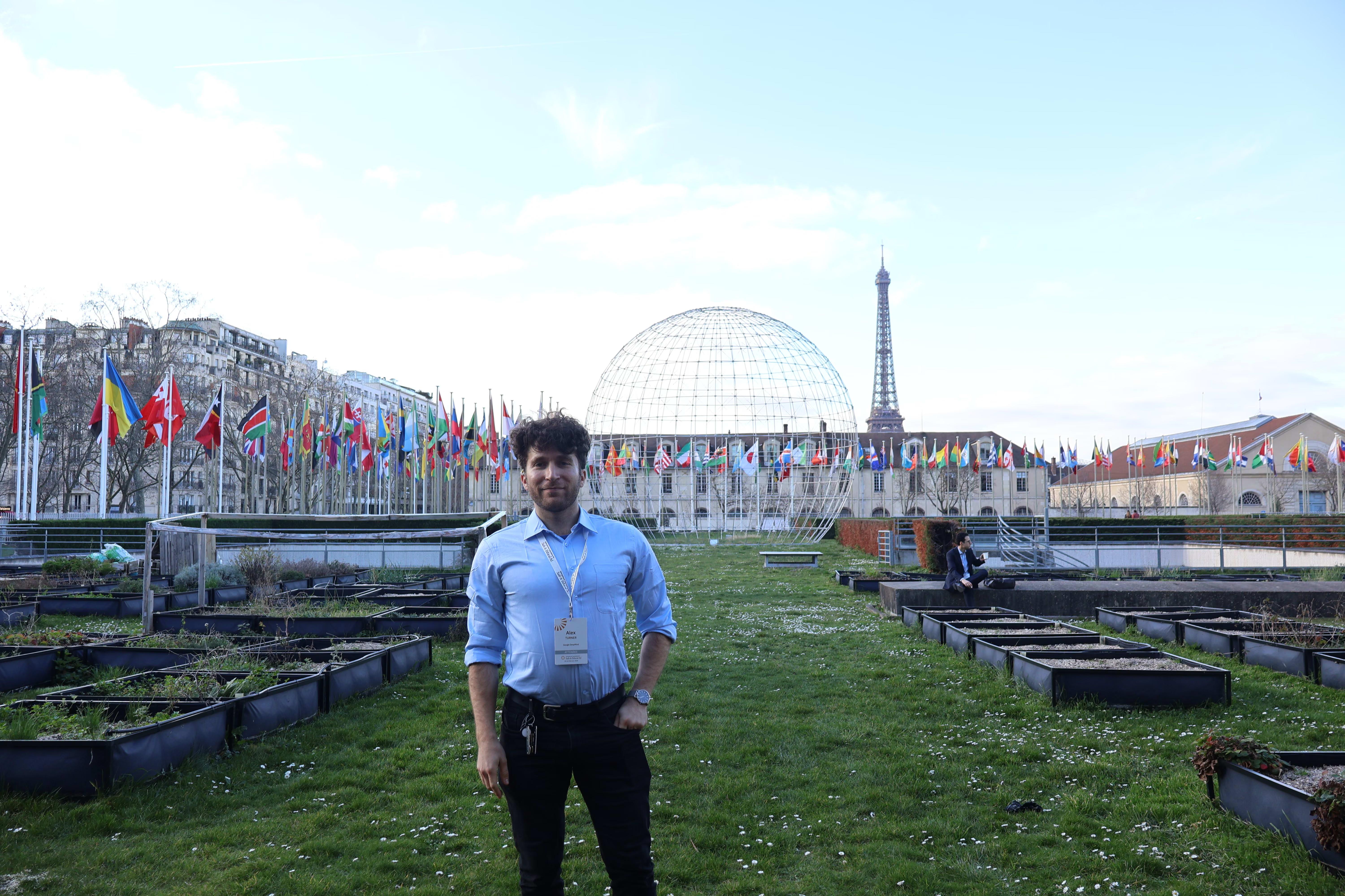 Alex Turner stands in a garden at the UNESCO headquarters in Paris, wearing a conference badge. In the background, a large metal dome structure and numerous international flags are visible, with the Eiffel Tower rising in the distance.
