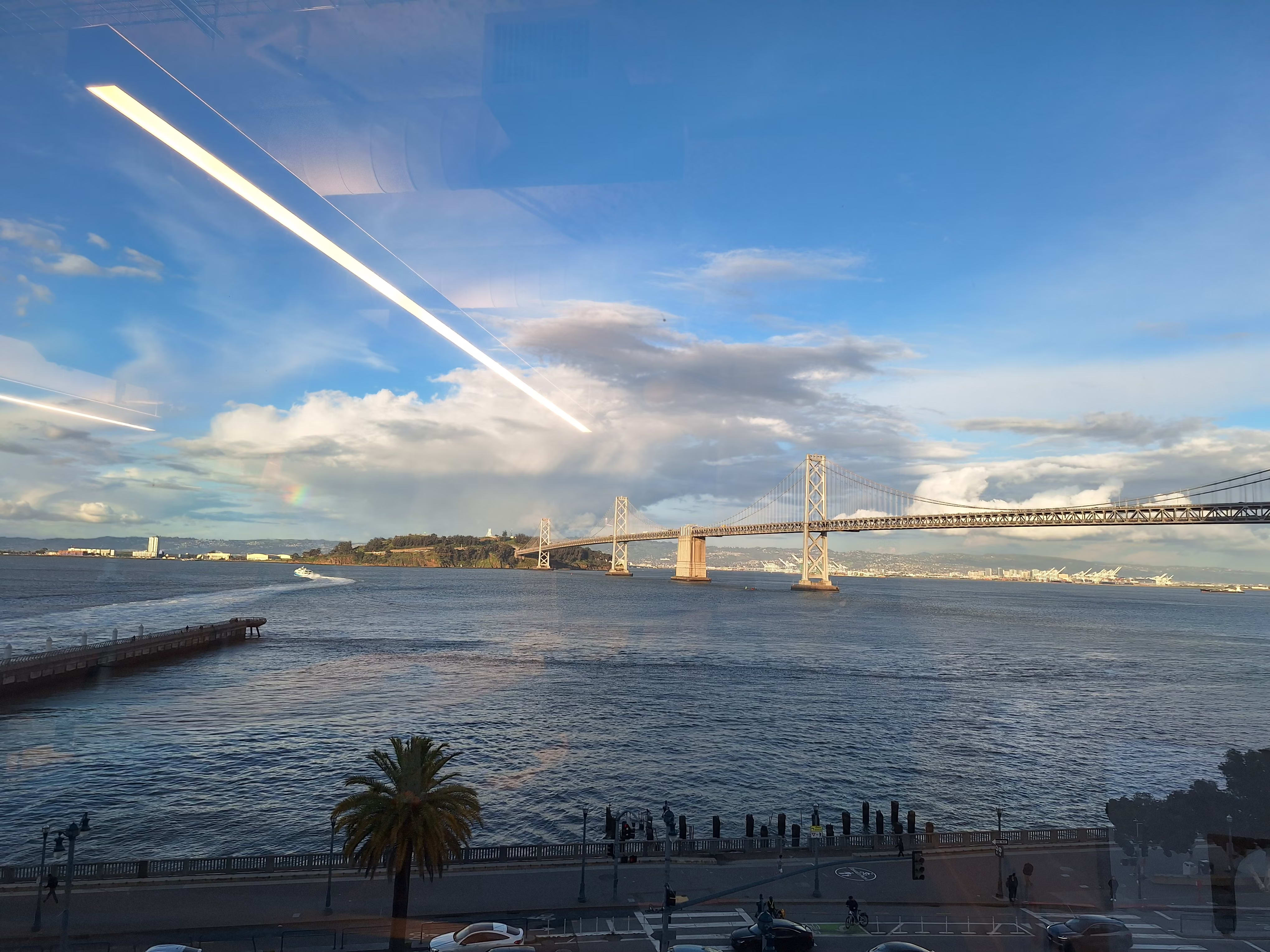 The view from an office window overlooking the San Francisco–Oakland Bay Bridge, showing the blue water of the bay, a palm tree in the foreground, and a coastal road with traffic below. A small section of rainbow peeks above Treasure Island.