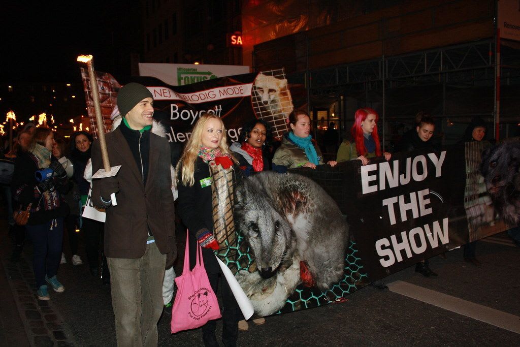 A nighttime street protest march against the fur fashion industry. Demonstrators carry torches and banners, including one reading "NEJ TIL BLODIG MODE / Boycott [Valenti...]" (Danish for "No to Bloody Fashion / Boycott...") and another large black banner displaying a graphic image of a skinned or injured animal alongside the text "ENJOY THE SHOW."